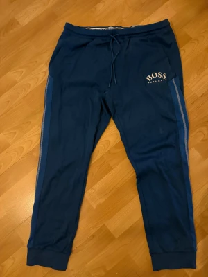 Blå mjukisbyxor från Hugo Boss - Snygga blå mjukisbyxor från Hugo Boss med logga på vänster sida. Byxorna har snörning i midjan och ribbade muddar vid bensluten. Snygga blå ränder längs sidorna ger en sportig vibe. Perfekta för chill eller träning.