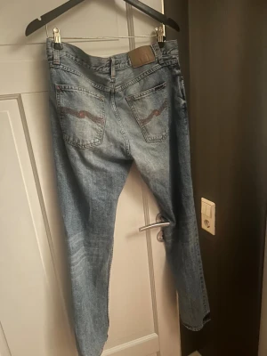 Nudie Jeans ljusblå jeans 31/32 - Säljer ett par ljusblå jeans från Nudie Jeans i klassisk femficksmodell. Jeansen har orangea sömmar, Nudie-logga på bakfickorna och läderpatch i midjan. Passformen är rak och normal, tillverkade i bomull. Perfekt för dig som gillar stilrena och tidlösa jeans.