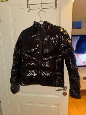 Svart glansig pufferjacka från Colmar - Säljer en svart, glansig pufferjacka från Colmar med klassisk logga på ärmen. Jackan har hög krage, dragkedja framtill och quiltad design. Perfekt för kalla dagar och ger en riktigt clean look. Materialet är syntet och jackan är långärmad. Det är storlek 42 men det är italienska storlekar då det är 36 i Svensk storlek