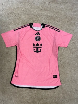 Inter Miami rosa fotbollströja Adidas - Snygg rosa Inter Miami fotbollströja från Adidas med svarta detaljer och klubbmärke på bröstet. Tröjan har korta ärmar, svarta ränder på axlarna och svart logga framtill. Tillverkad i lätt och ventilerande funktionsmaterial.