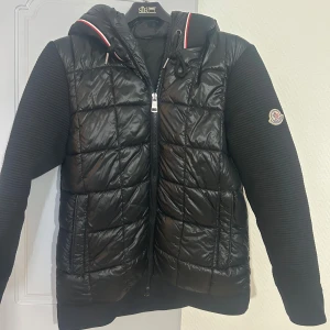 Moncler Cardigan - Moncler Cardigan i storlek S med quiltad framsida och stickade ärmar. Jackan har huva med broderad logga och rödvit kant, dragkedja framtill och Moncler-märke på ärmen. Perfekt för dig som vill ha en snygg och stilig stil?