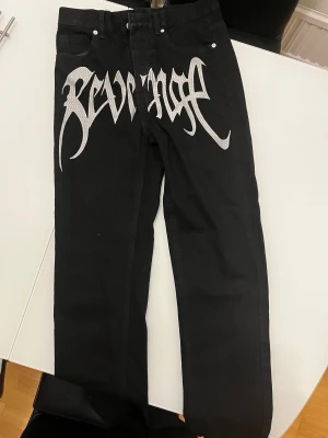 Svarta jeans Revenge - Unika svarta jeans med stort silvrigt Revenge-tryck över framsidan. Klassisk femficksmodell med raka ben och bälteshällor. Jeansen har en cool streetwear-vibe och är tillverkade i slitstarkt denim-material.