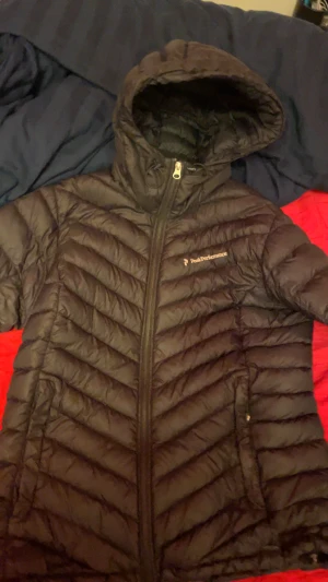 Svart dunjacka Peak Performance - Svart dunjacka från Peak Performance med huva och snyggt quiltad design. Jackan har dragkedja framtill och diskret logga på bröstet. Perfekt för kalla dagar med sin lätta och värmande känsla. Passar dig som gillar stilren och sportig look.