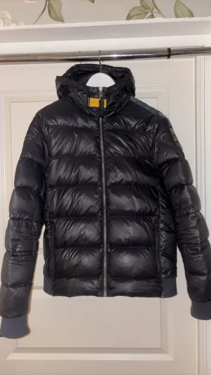 Svart dunjacka från Parajumpers - Svart, quiltad dunjacka från Parajumpers med huva och ribbade muddar. Jackan har en snygg patch på ärmen och dragkedja framtill. Perfekt för kalla dagar och har en cool, sportig vibe. Passformen är normal och materialet är glansigt syntet med dunfyllning.