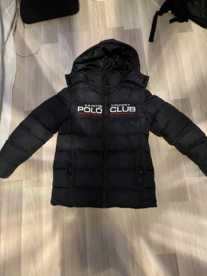 Svart pufferjacka från Polo Club - Svart pufferjacka från Polo Club Saint Vincent med huva och broderad logga på bröstet. Jackan har dragkedja framtill, två sidofickor med dragkedja och en badge på ärmen. Perfekt för kalla dagar med sin tjocka vaddering och stilrena look.