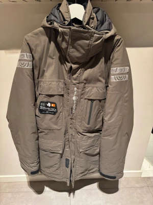 Oliv Grön Sail Racing parkajacka Gore-Tex - Grön färg med broderade loggor och patch 'Antarctica Expedition'. Jackan har flera fickor, huva och detaljer i silver. perfekt för kalla dagar. Snygga reflexdetaljer på ärmarna och dragkedja framtill.
