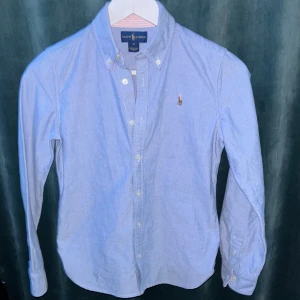 Blå skjorta från Ralph Lauren - Snygg ljusblå skjorta från Ralph Lauren med klassisk button-down krage och broderad logga på bröstet. Skjortan har långa ärmar, knappar framtill och är gjord i mjuk bomull. Perfekt för dig som gillar stilrena och tidlösa plagg. Använd en gång. Pris 399, kan diskuteras.
