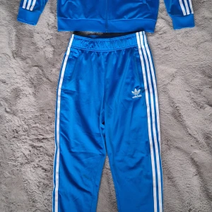 Adidasdress Adicolor SST Track set stl 158 - Stället är i mycket gott skick. Liten fläck från cykelkedja på ena benet, kolla bild 4.   Färg: Blue bird.  Normal passformSnygga blå träningsbyxor och träningsjacka från Adidas med klassiska vita ränder längs sidorna och Adidas-logga på låret. Byxorna har elastisk midja och är gjorda i glansigt syntetmaterial som ger en sportig vibe. Perfekta för chill eller träning.