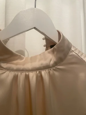 Champagnefärgad satinblus från Bik Bok - Säljer en elegant champagnefärgad blus från Bik Bok i storlek XS. Blusen är gjord i glansigt satinmaterial, har lång ärm, hög krage med knappar i nacken och ett lätt puffigt avslut vid midjan. Perfekt för dig som gillar stilrena och lyxiga vibes.