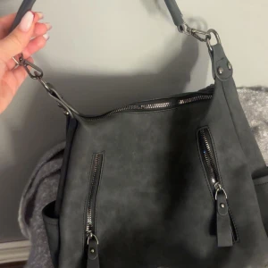 Svart axelväska i mockaimitation - Snygg svart axelväska i mockaimitation med flera dragkedjefickor framtill och baktill. Väskan har bred axelrem med metallspännen och rymligt foder inuti. Perfekt för dig som vill ha en stilren och praktisk väska med coola detaljer. Fick denna i födelsedagspresent därav vet jag ej nypris eller vart den kommer ifrån.