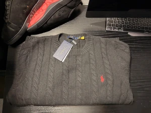 Svart Polo Ralph Lauren - Svart kabelstickad tröja från Polo Ralph Lauren med klassisk röd broderad logga på bröstet. Tröjan har rund halsringning, ribbade muddar och är tillverkad i mjukt material. Perfekt för dig som gillar stilren och tidlös design.