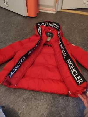 Röd Moncler pufferjacka med huva - ❌ använd 1 gång växte ur  innan han,  hann börja använda den.   Säljer denna pufferjacka från Moncler med stor huva och tydlig logga på ärmen. Jackan har svarta detaljer med Moncler-tryck längs dragkedjan och huvan, samt två stooora  sidofickor med dragkedja. Perfekt för kalla dagar och riktigt snygg streetstil.