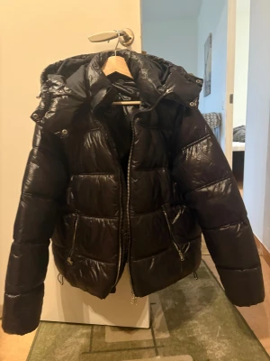 Svart pufferjacka med huva - Säljer en svart pufferjacka med stor huva och dragkedja framtill. Jackan har två fickor med dragkedjor och justerbar nederkant. Perfekt för kalla dagar och riktigt skön att bära. Materialet är glansigt och ger en modern look.