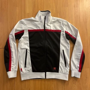 DC Track Jacket svart/vit/röd - Snygg track jacket från DC med svart front och rygg, vita ärmar och sidor samt röda detaljer längs bröst och armar. Jackan har hög krage, dragkedja framtill och två fickor med dragkedja. Perfekt för en sportig och street-inspirerad look. 