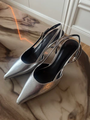 Silvriga klackskor med spetsig tå från H&M - Säljer ett par silvriga klackskorfrån H&M med smal klack och spetsig tå, jag säljer de då jag inte fått någon användning av de. Skorna har öppen häl med justerbar rem och en glansig metallic-finish. Perfekta för att lyfta vilken outfit som helst!!!! 