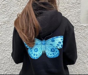 Svart hoodie med blå fjäril - Svart hoodie med stor blå fjäril på ryggen. Tröjan har huva och en relaxed passform, perfekt för dig som gillar statement-prints. Materialet känns mjukt och skönt, och den har långa ärmar. Snygg detalj med den färgstarka fjärilen som verkligen poppar mot det svarta tyget.