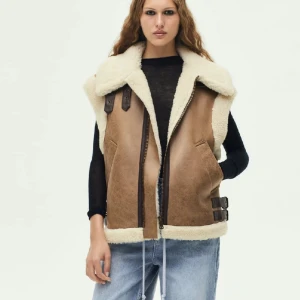 Beige pälsväst från Zara med detaljer - Säljer en beige pälsväst från Zara med fluffig vit fuskpäls på insidan och krage. Västen har två fickor, bruna spänndetaljer på sidorna och dragkedja framtill. Perfekt för lager-på-lager och har en loose passform med coola detaljer.