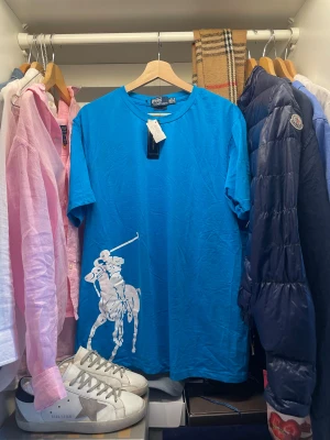 Blå t-shirt Polo Ralph Lauren XXL - Snygg blå t-shirt från Polo Ralph Lauren i storlek XXL. T-shirten har en stor vit polospelare tryckt framtill. Tröjan kostar runt 1300 ny. Nytt skick med pristag och är aldrig använd. Perfekt för vardagen.                                                                           Skriv vid frågor, priset är inte hugget i sten🤝