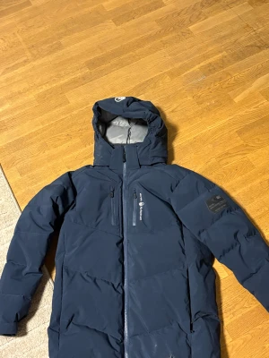 Mörkblå Sail Racing dunjacka - Säljer en mörkblå dunjacka från Sail Racing, modell Antarctica Expedition. Jackan har huva, flera praktiska fickor med dragkedja och snygga Sail Racing-loggor på bröstet och ärmen. Perfekt för kalla dagar med sin quiltade design och stilrena look.