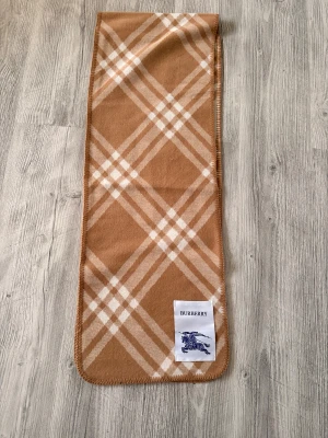 Burberry Halsduk - Snygg halsduk från Burberry i klassiskt rutigt mönster med beige och vita toner. Halsduken har en rektangulär form och är mjuk med en stor Burberry-logga i ena hörnet. Perfekt för vintern!