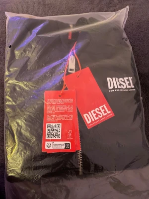 Svart hoodie med dragkedja från Diesel. 12 year barn - Svart hoodie från Diesel med vit logotyp på bröstet och röd etikett i nacken. Tröjan har huva, hel dragkedja framtill och två fickor. Tillverkad i mjuk bomull, perfekt för chill dagar. Klassisk och stilren look med ribbade muddar vid ärmslut och nederkant.