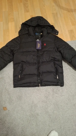 Svart pufferjacka från Polo Ralph Lauren - Svart pufferjacka från Polo Ralph Lauren med huva och två dragkedjeförsedda fickor. Jackan har det klassiska röda Polo-logot broderat på bröstet och en snygg quiltad design. Perfekt för kalla dagar och har en modern, clean look.