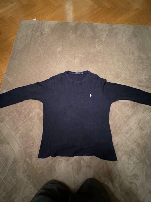Mörkblå långärmad tröja Polo Ralph Lauren - Snygg mörkblå långärmad tröja från Polo Ralph Lauren med klassisk rund hals och vit broderad logga på bröstet. Tröjan är ribbad och har en skön passform, perfekt för dig som gillar stilrena och tidlösa plagg. (🔥om det syns några fleckar så är det sånt som går att tvätta 🧽 🔥)