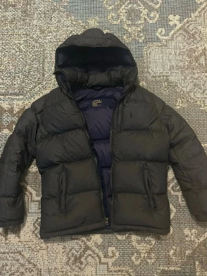 Svart pufferjacka från Polo Ralph Lauren - Säljer en svart pufferjacka från Polo Ralph Lauren med huva och dragkedja. Jackan har två sidofickor och diskret logga på bröstet. Perfekt för kalla dagar och riktigt skön att bära. Passar dig som gillar klassisk och sportig stil.
