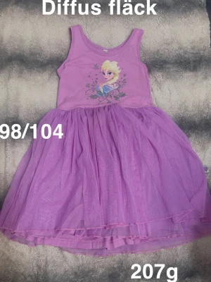 Lila Frozen-klänning med tyllkjol - Söt lila klänning med motiv av Elsa från Frozen på bröstet och fluffig tyllkjol. Ärmlös modell med rund halsringning och voluminös kjol som ger prinsesskänsla. Perfekt för dig som älskar Disney och vill ha något extra fint.🌸 Diffus fläck. Kan finnas någon dragning i tyllen som det blir direkt. Storlek 98/104