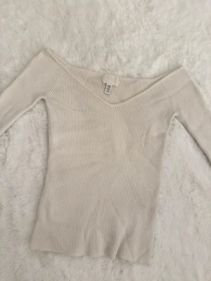 Offshoulder ribbad topp  - Superfin offshoulder topp från H&M i ljusbeige färg. Toppen är ribbad och har lång ärm med en snygg v-ringning. Tillverkad i mjuk bomull som sitter skönt och följer kroppen. Perfekt att styla med jeans eller kjol för en trendig look. Helt ny.