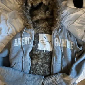 Grå zip up från Abercrombie & Fitch - Mysig grå hoodie med pälskantad huva från Abercrombie & Fitch. Jackan har dragkedja och stora tryckta bokstäver på framsidan. Perfekt för kyliga dagar med sin fluffiga insida.💗
