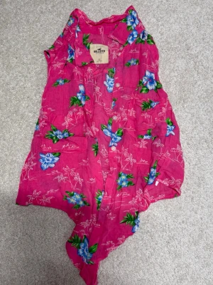 Blommig rosa blus från Hollister - Säljer en ärmlös blus från Hollister i stark rosa med blå och gröna blommor. Blusen har krage, knappar framtill och knytning nedtill för en chill sommarlook. Materialet känns lätt och luftigt, perfekt för varma dagar.