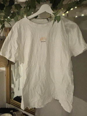 Vit t-shirt med broderi från 157 - Vit t-shirt från 157 i 100% bomull. Framtill finns ett litet motiv av en sol och texten 'SUN'S OUT' i svart. Klassisk rund hals och korta ärmar, perfekt till sommaren. Enkel och clean design som passar till allt.