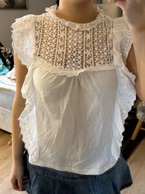 Vit blus med spets och volanger - Säljer en superfin vit blus med detaljerad spets upptill och volanger längs axlar och sidor. Blusen har hög hals med liten volangkant och är ärmlös. Perfekt för dig som gillar romantisk och söt stil. Materialet känns lätt och luftigt.