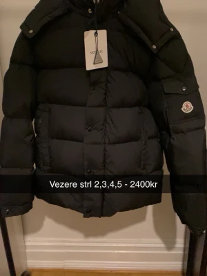 Svart pufferjacka från Moncler - Säljer en svart pufferjacka från Moncler med klassisk logga på ärmen och hög krage. Jackan har knappar och dragkedja framtill samt fickor på sidorna. Perfekt för kalla dagar och riktigt snygg till alla outfits.