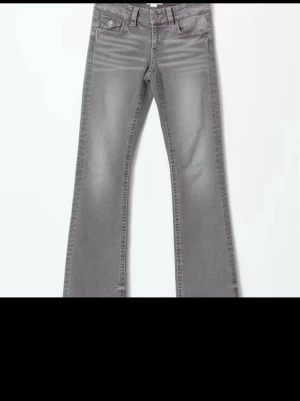 Grå bootcut jeans med fickdetaljer - Snygga grå jeans med bootcut passform och coola fickor med lock och knapp bak. Jeansen har en klassisk femficksdesign, bälteshällor och är tillverkade i mjukt jeansmaterial. Perfekta till sneakers eller boots för en avslappnad look.