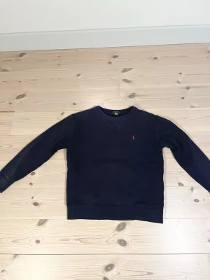 Mörkblå sweatshirt från Polo Ralph Lauren - Mörkblå fleece sweatshirt från Polo Ralph Lauren med klassisk röd logga broderad på bröstet. Skick 7/10 då ärmarna på tröjan är slitna. killen på bilden är 157cm. Storlek 150. Nypris ca 1200kr