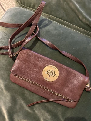 Vintage brun crossbody foldover  Mulberry-väska - Supersnygg vintage brun axelväska från Mulberry. Väskan har ett stort guldfärgat Mulberry-emblem framtill som tyvärr är lite repigt (kan skicka bättre bild). Justerbar axelrem som bärs som crossbody, och kan sättas fast på två olika ställen, om man inte vill att väskan ska vara foldover, och därmed blir dubbelt så stor. Två extra fickor på utsidan som göms under foldovern. Vintage från 00-talet, därmed en del slitage. 