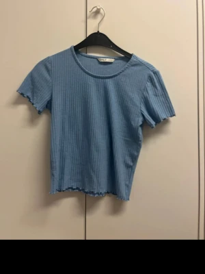 Blå ribbad topp från Only - Säljer en ljusblå ribbad topp från Only med korta ärmar och vågig kant vid ärmslut och nederkant. Toppen har rund halsringning och är i mjukt, stretchigt material som sitter snyggt på kroppen.