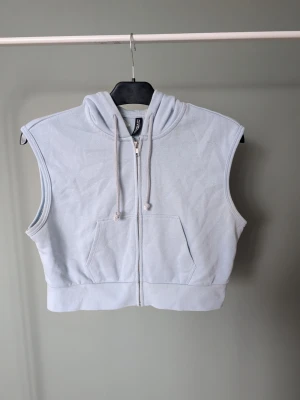 Ljusblå hoodie-väst - Säljer en ljusblå hoodie-väst från H&M Divided. Västen har dragkedja framtill, huva med snören och en stor magficka. Perfekt för lager på lager och chill stil. Materialet är mjuk bomull som känns skönt mot huden.