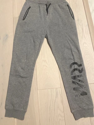 Grå mjukisbyxor med tryck - Säljer ett par grå mjukisbyxor från stadium med svart tryck på ena benet. Byxorna har snörning, dragkedjeförsedda fickor fram och en bakficka med liten logga. perfekta för chill eller träning.