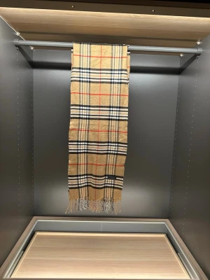 Halsduk från Burberrys - Säljer en klassisk rutig väst från Burberrys i beige, svart, vit och röd. Västen är tillverkad i 100% lammull och har fransar längst ner. Perfekt för dig som vill ha en tidlös och ikonisk look.