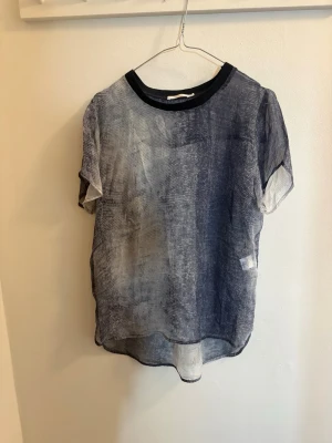 Blå transparent blus med svart krage - Säljer en luftig och transparent blus i blått och vitt med subtilt mönster. Blusen har korta ärmar och en svart rund krage. Materialet är tunt och lätt, perfekt för varmare dagar eller lager-på-lager. Passar dig som gillar en avslappnad men cool stil.