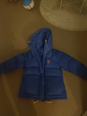 Blå dunjacka från Fjällräven aldrig använt köpte för nån vecka sne men ångar mig med färgen därför säljer jag den , då jag dragit av lappen så de går inte att lämna tillbaka  - Säljer en blå dunjacka från Fjällräven med huva och gula detaljer. Jackan har stora fickor framtill och klassisk Fjällräven-logga på bröstet. Perfekt för kalla dagar och riktigt skön att bära. HELT NYY!!!!!!!