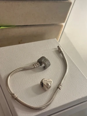 Smyckesarmband med berlocker från Pandora - Säljer ett stilrent silverfärgat armband från Pandora med två berlocker. Ena berlocken är hjärtformad med texten 'Sister' och en liten sten, den andra är rund och glittrig med små stenar. Perfekt accessoar för att lyfta din look. Armbandet har gått ur sortimentet vilket innebär att detta pieace är (vintage) då den inte finns att köpa längre. 