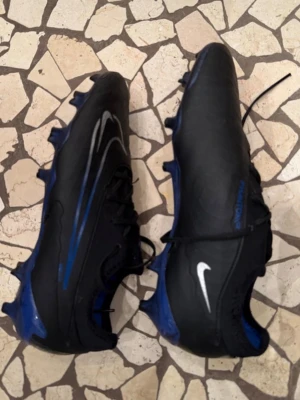 Nike svarta och blåa fotbollsskor - Säljer ett par svarta och blåa Nike fotbollsskor med vita detaljer och snörning. Skorna har dobbar för bra grepp på planen och en sportig design med rund tå. Perfekta för dig som vill ta ditt spel till nästa nivå. Priset kan diskuteras.