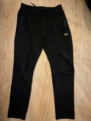 Svarta mjukisbyxor från Lacoste - Svarta mjukisbyxor från Lacoste med klassisk krokodillogo på vänster sida. Byxorna har dragsko i midjan, två sidofickor och en bakficka. Tillverkade i mjukt material som passar perfekt för chill dagar. Enkel och stilren design.