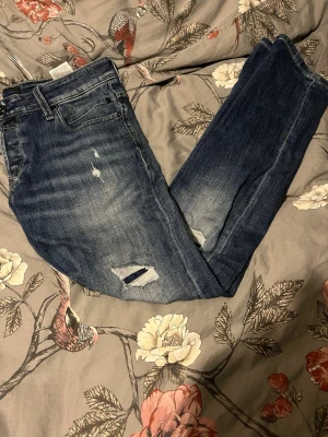 Blå slitna jeans från Jack & Jones - Säljer ett par blå jeans från Jack & Jones med slitningar och hål på benen för en riktigt avslappnad look. Jeansen har klassisk femficksdesign, normal passform och är tillverkade i mjukt denimtyg. Perfekta för dig som gillar en casual och trendig stil.