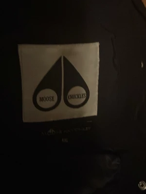 Svart Moose Knuckles dunjacka XXL - Säljer en svart dunjacka från Moose Knuckles i storlek M.Jackan har en stor huva med vit päls, silverfärgade dragkedjor och knappar samt logga på ärmen. Perfekt för kalla vinterdagar och riktigt snygg streetstil.