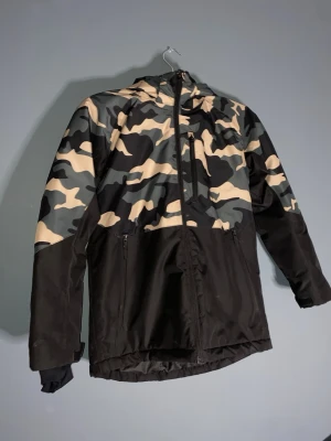 Camouflage dunjacka från Everest - Säljer en snygg dunjacka från Everest i storlek 158. Jackan har ett grått, svart och beige camouflage-mönster upptill och svart nedtill. Utrustad med huva, dragkedjor och flera fickor. Perfekt för dig som gillar streetwear och vill sticka ut.